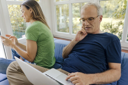 Mature couple sitting on couch at home with man using laptop and woman using cell phone - KNSF04644