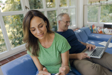 Mature couple sitting on couch at home with woman using cell phone and man using laptop - KNSF04645