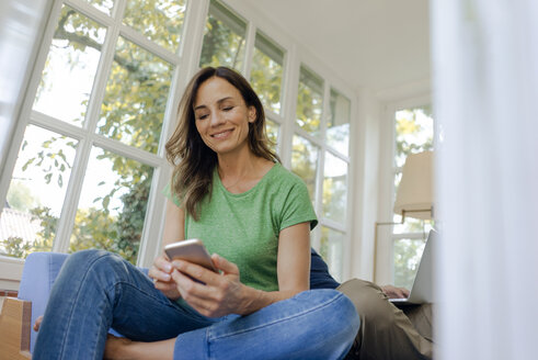 Smilong mature woman sitting on couch at home using cell phone with man in background - KNSF04646
