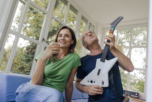 Mature couple sitting on couch at home with man playing toy electric guitar - KNSF04648