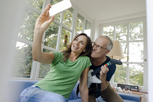 Happy mature couple taking a selfie at home with man playing toy electric guitar - KNSF04650