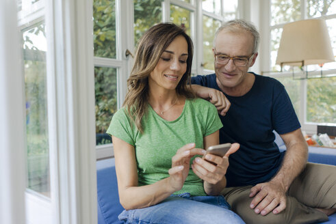 Smiling mature couple sitting on couch at home sharing cell phone - KNSF04652