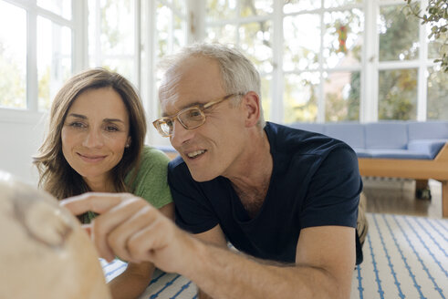 Mature couple lying on the floor at home looking at globe - KNSF04654