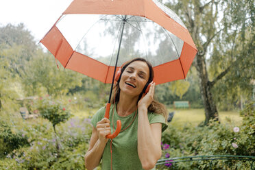 Glückliche reife Frau steht im Regen unter Regenschirm und hört Musik mit Kopfhörern - KNSF04665