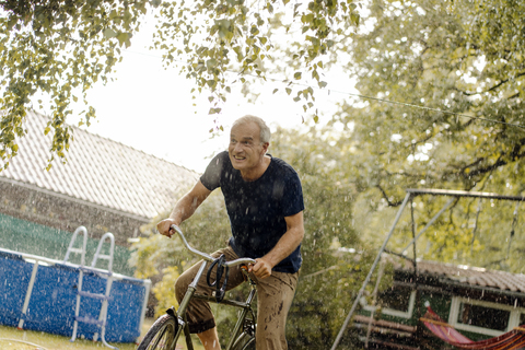 Glücklicher reifer Mann fährt Fahrrad im Sommerregen im Garten, lizenzfreies Stockfoto