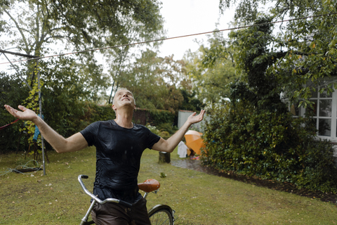 Mature man with bicycle enjoying summer rain in garden stock photo