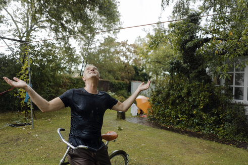 Älterer Mann mit Fahrrad genießt den Sommerregen im Garten - KNSF04672