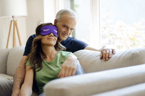 Lächelndes reifes Paar sitzt zu Hause auf der Couch, die Frau trägt eine Augenmaske, lizenzfreies Stockfoto