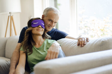 Lächelndes reifes Paar sitzt zu Hause auf der Couch, die Frau trägt eine Augenmaske - KNSF04707