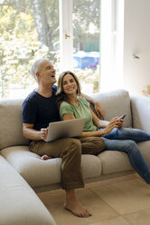 Happy mature couple sitting on couch at home with cell phone and laptop - KNSF04708