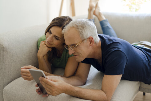 Lächelndes reifes Paar, das zu Hause auf der Couch liegt und ein Tablet teilt - KNSF04709