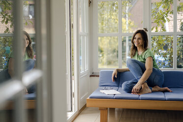 Smiling mature woman sitting on couch at home with book - KNSF04710