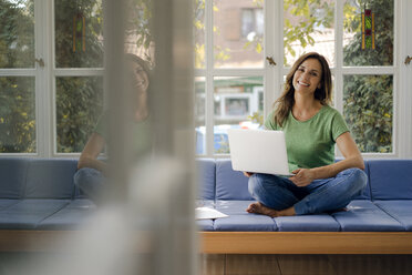 Smiling mature woman sitting on couch at home with laptop - KNSF04712