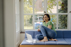 Lächelnde reife Frau sitzt auf der Couch zu Hause mit Laptop - KNSF04717