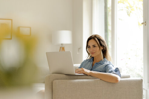 Mature woman sitting on couch at home using laptop - KNSF04734