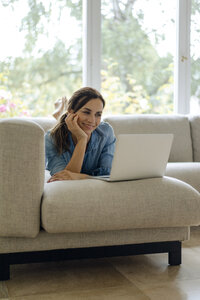 Lächelnde reife Frau auf der Couch liegend zu Hause mit Laptop - KNSF04736