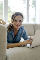 Portrait of smiling mature woman lying on couch at home using laptop - KNSF04738