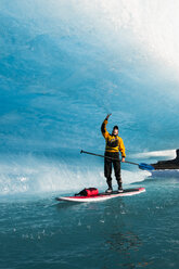 Ein Mann auf einem Stand Up Paddle Board (SUP) berührt eine Welle aus Eis, die auf dem Bear Lake im Kenai Fjords National Park, Alaska, abtropft. - AURF02691