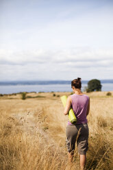 Eine junge Frau in einem lila Hemd trägt eine grüne Yogamatte durch ein Feld mit goldenen Gräsern und Blick auf den Ozean. - AURF02935