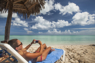 Eine junge Frau macht ein Foto mit ihrem Smartphone, während sie sich am Strand entspannt - AURF02974