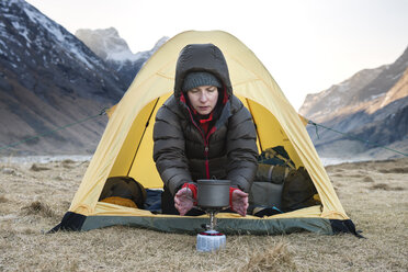 Female hiker warms hands with stove on cold morning wild camping at Horseid beach, Moskenes├©y, Lofoten Islands, Norway - AURF03115