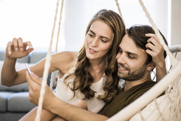 Couple in hanging chair at home looking at tablet - JOSF02552
