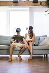 Couple sitting on couch at home wearing VR glasses playing video game - JOSF02563