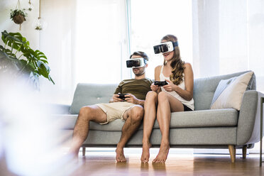 Couple sitting on couch at home wearing VR glasses playing video game - JOSF02564