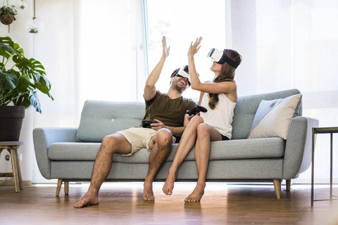 Glückliches Paar, das zu Hause auf der Couch sitzt und eine VR-Brille trägt, High Fiving, lizenzfreies Stockfoto