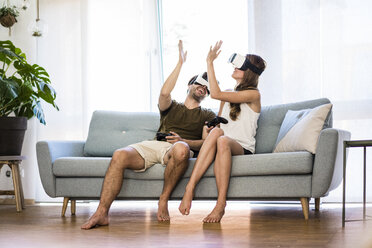 Happy couple sitting on couch at home wearing VR glasses high fiving - JOSF02566