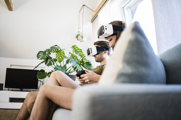 Couple sitting on couch at home wearing VR glasses playing video game - JOSF02633