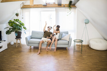 Happy couple sitting on couch at home wearing VR glasses high fiving - JOSF02634