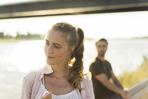 Frau isst Eis im Sommer am Flussufer mit Mann im Hintergrund, lizenzfreies Stockfoto