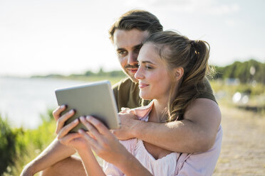 Smiling couple at the riverside in summer holding tablet - JOSF02666