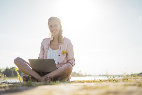 Porträt einer lächelnden Frau, die im Sommer am Flussufer sitzt und einen Laptop benutzt - JOSF02668