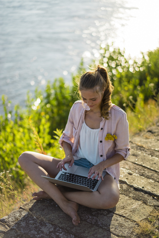 Frau sitzt im Sommer am Flussufer und benutzt einen Laptop, lizenzfreies Stockfoto