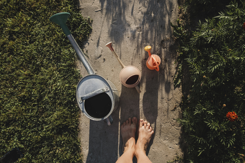 Drei Gießkannen und die Füße einer Frau auf einem Gartenweg, Draufsicht, lizenzfreies Stockfoto