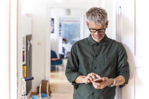 Mature man leaning against door case at home using cell phone - TCF05814