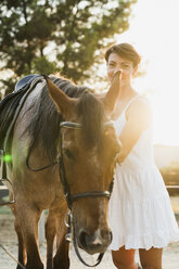 Portrait of smiling woman standing besides riding horse at backlight - KKAF01588