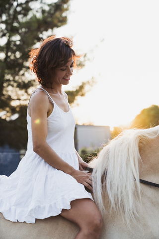 Lächelnde Frau in weißem Kleid reitet ohne Sattel auf einem Pferd bei Sonnenuntergang, lizenzfreies Stockfoto