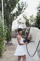 Porträt einer glücklichen Frau im weißen Kleid, die ein Pferd führt - KKAF01619