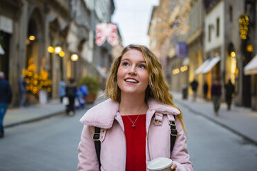 Italien, Florenz, Porträt eines lächelnden Touristen, der mit einem Kaffee zum Mitnehmen auf der Straße spazieren geht - MGIF00243