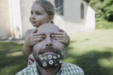 Little girl's hands covering eyes of mature man with daisies in his beard - KMKF00468