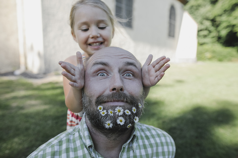 Porträt eines reifen Mannes mit Gänseblümchen in seinem Bart, der mit seiner kleinen Tochter im Garten spielt, lizenzfreies Stockfoto