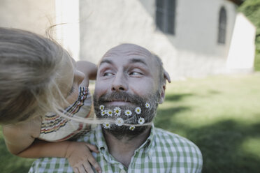 Porträt eines lächelnden reifen Mannes mit Gänseblümchen in seinem Bart, der mit seiner kleinen Tochter im Garten spielt - KMKF00470