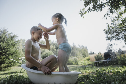 Brother and sister playing with water in little bath tub in garden - KMKF00494