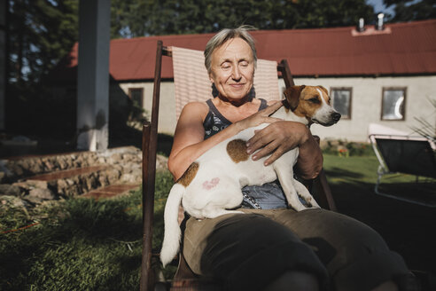 Smiling senior woman with dog on deckchair in garden - KMKF00509