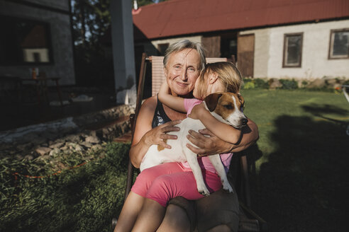 Smiling grandmother with granddaughter and dog on deckchair in garden - KMKF00510