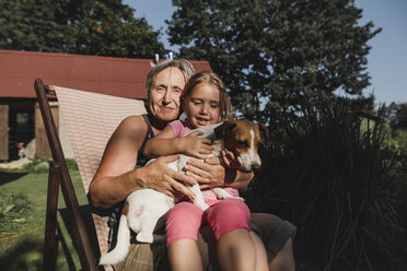 Lächelnde Großmutter mit Enkelin und Hund auf einem Liegestuhl im Garten - KMKF00511