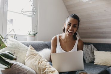 Porträt einer lachenden Frau, die mit ihrem Laptop zu Hause auf der Couch sitzt - LHPF00004